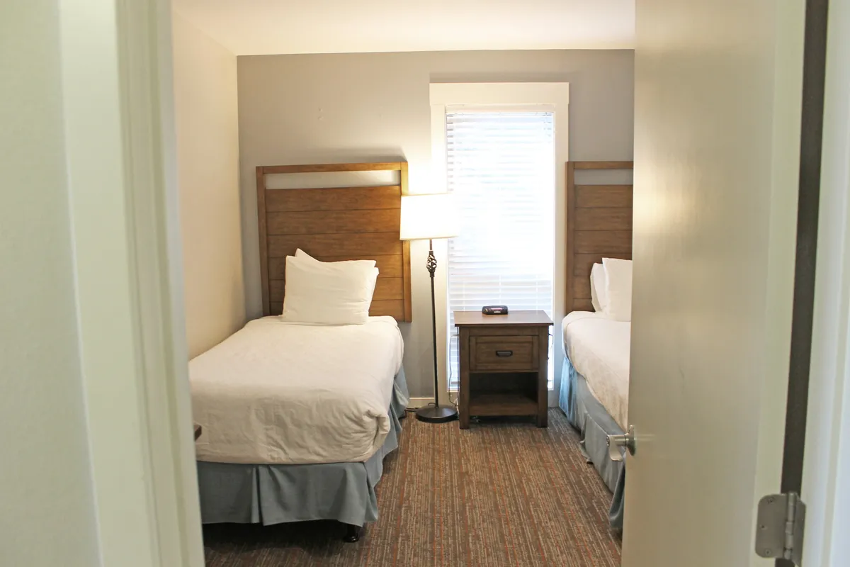 This image displays a cozy bedroom with two twin beds separated by a nightstand and lamp. The bedding is neatly arranged and the space feels bright and welcoming, ideal for both children and adult pairs.