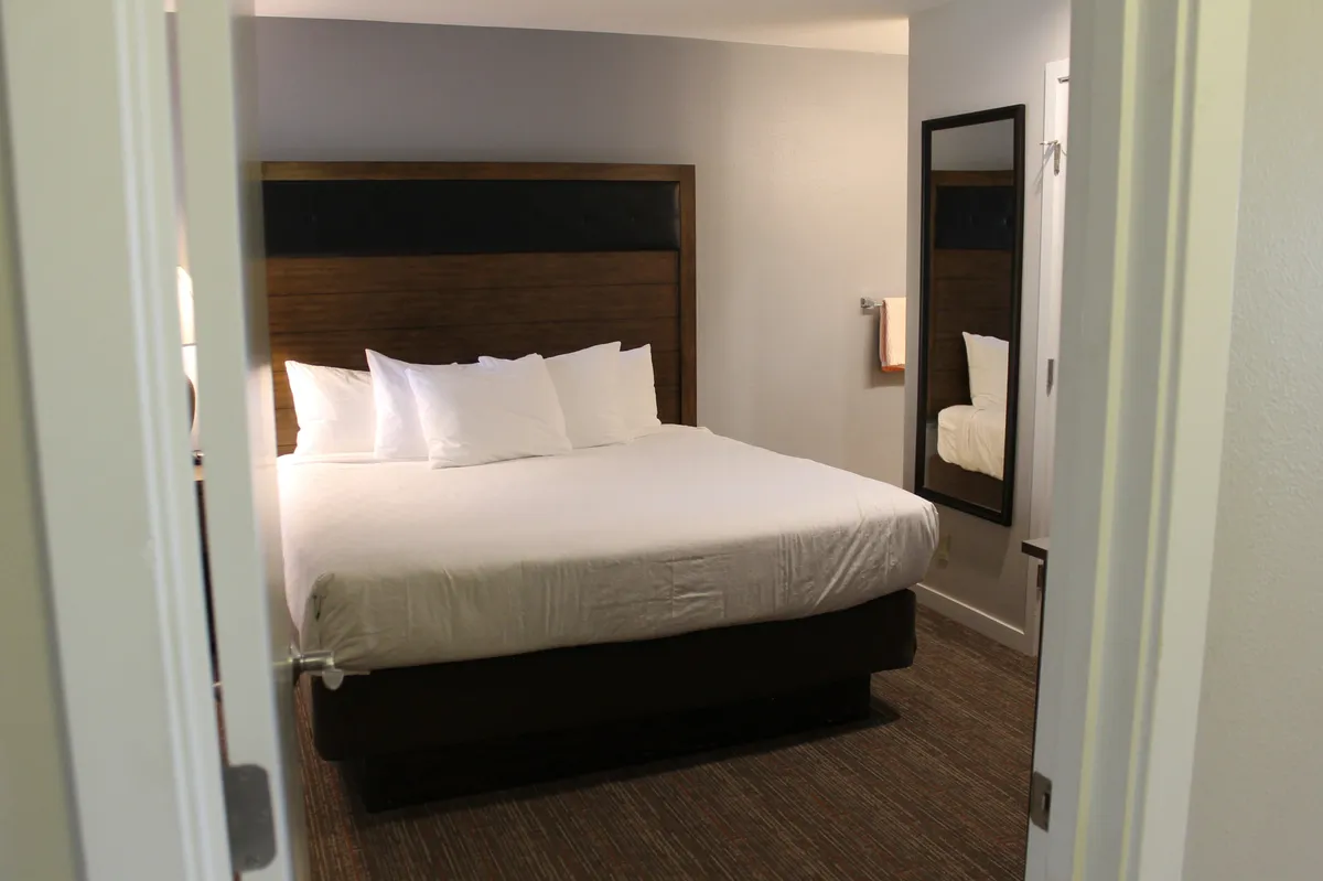 This image shows a serene primary bedroom featuring a king-sized bed with elegant white bedding, a wooden headboard, and a full-length mirror. The space is softly lit, evoking a restful and inviting ambiance perfect for relaxation.