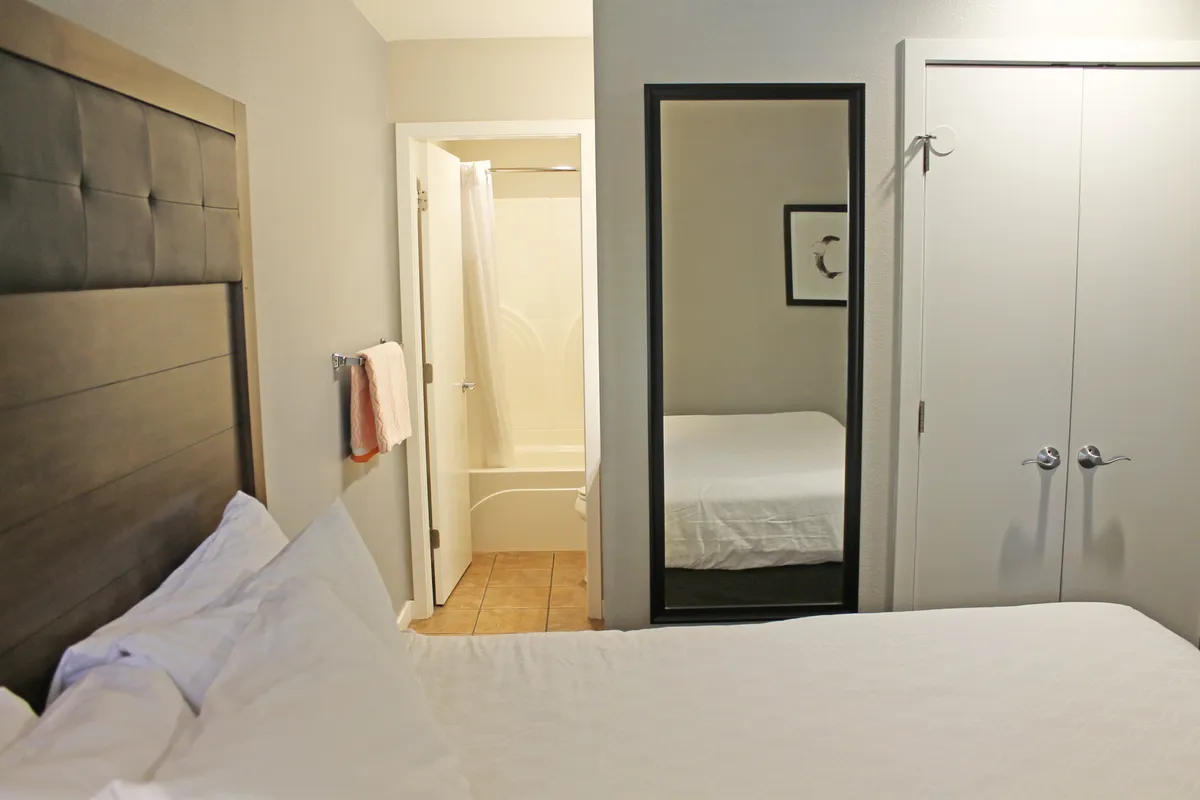 A comfortable queen bedroom is shown, complete with a large bed, a mirrored wardrobe, and the door leading to a private ensuite bath. The window in the background hints at pleasant views, enhancing the room’s sense of tranquility and light.