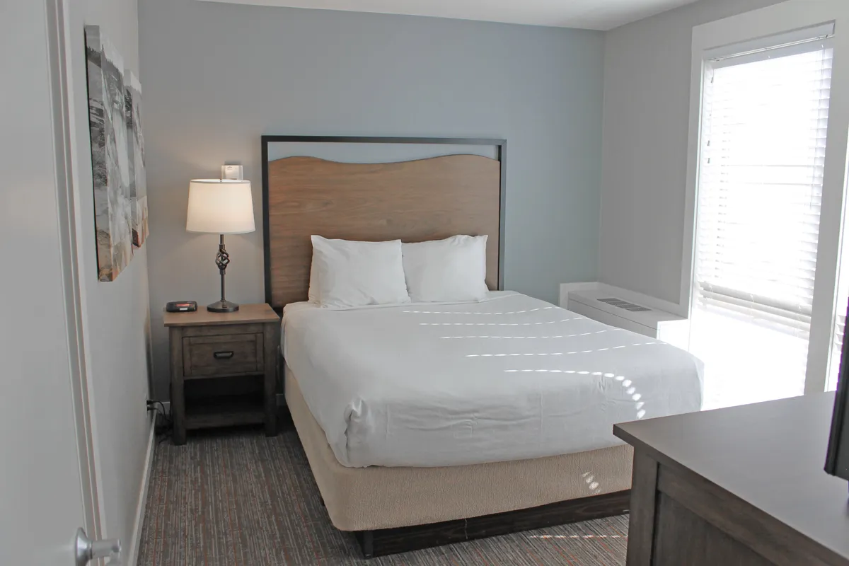 A tranquil bedroom with a neatly made bed, a nightstand, a lamp, and a window letting in natural light. The room is peaceful and inviting, suggesting restful nights and private retreat.