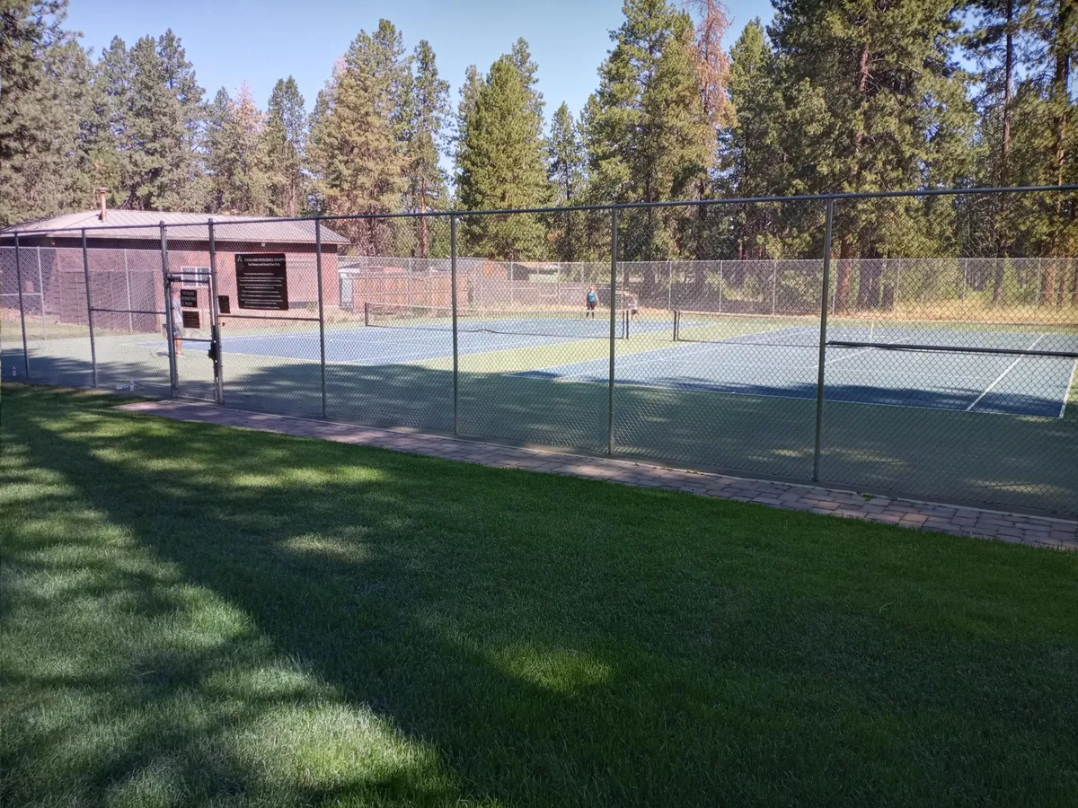 This image features well-maintained tennis courts bordered by grassy areas and trees, with a building visible in the background. The scene is lively yet relaxed, suggesting both active play and opportunities for relaxing nearby.
