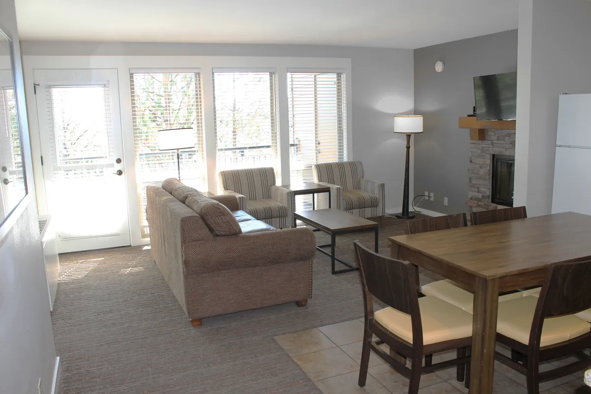 This image captures the main living area with a plush sofa, a dining table, a warm fireplace, and large windows bringing in abundant natural light. The space feels open and inviting, with the living, dining, and relaxing features all in view.