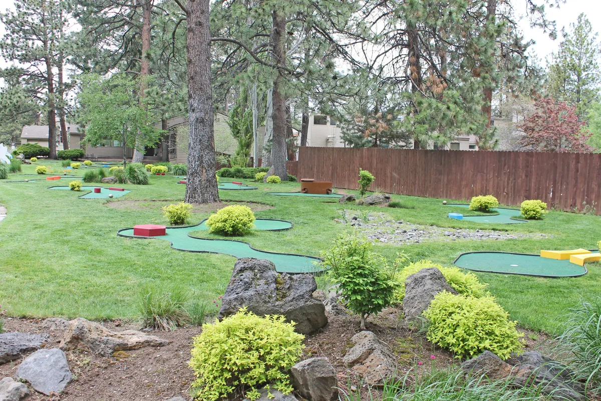 This image features a cheerful mini-golf course surrounded by lush greenery, decorative rocks, benches, and a wooden fence. The setting is vibrant and welcoming, with ample space for families to engage in friendly competitions and relax outdoors.