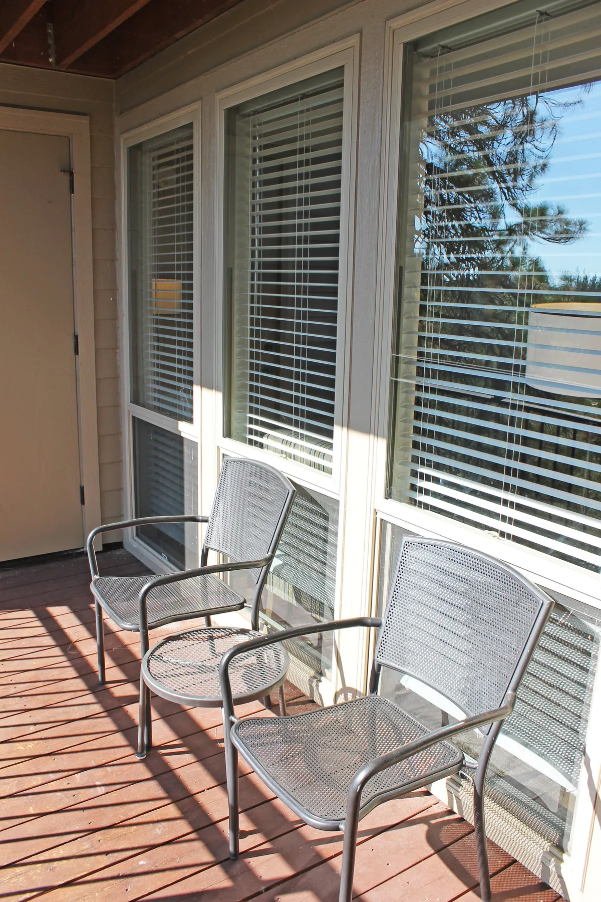 The image shows a serene wooden deck with two metal chairs overlooking a tranquil outdoor space. The sliding glass doors connect the interior living area with the outdoors, creating a seamless spot for relaxation, morning coffee, or shared moments as a group.